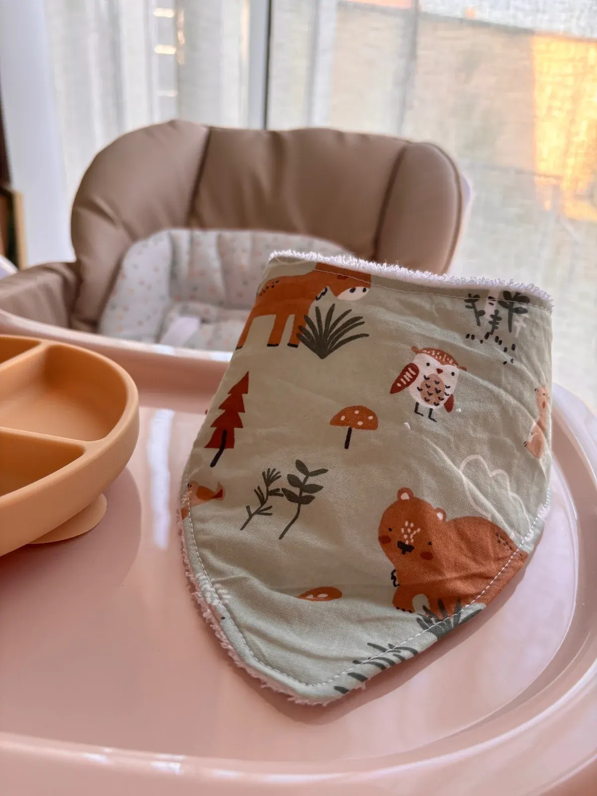 Bavoir bandana vert d'eau à motifs animaux de la forêt, posé sur la tablette d'une chaise haute.