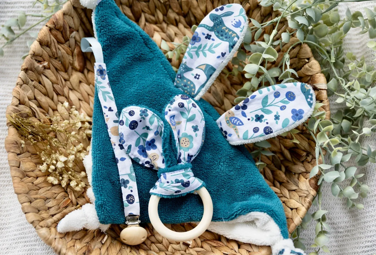 Doudou plat bleu canard, anneau de dentition et attache-tétine disposés dans une corbeille tressée