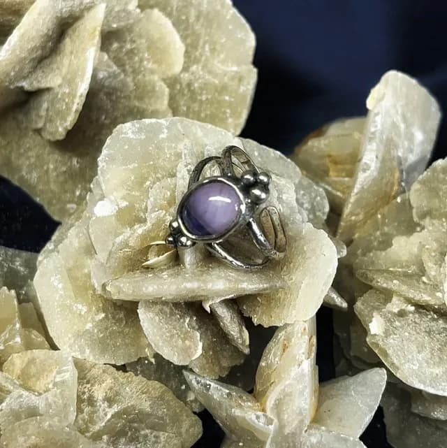 Bague en argent ornée d'un cabochon ovale violet, posée sur une rose des sables.