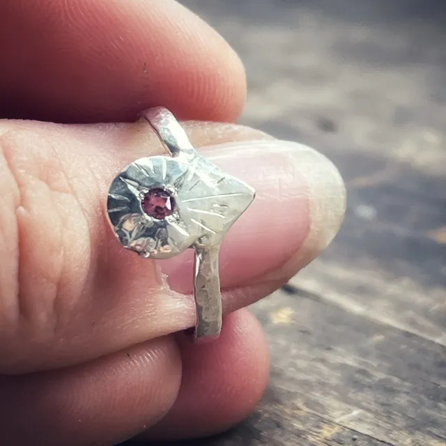 Gros plan sur le motif gravé et la petite pierre rose de la bague en argent tenue en main.