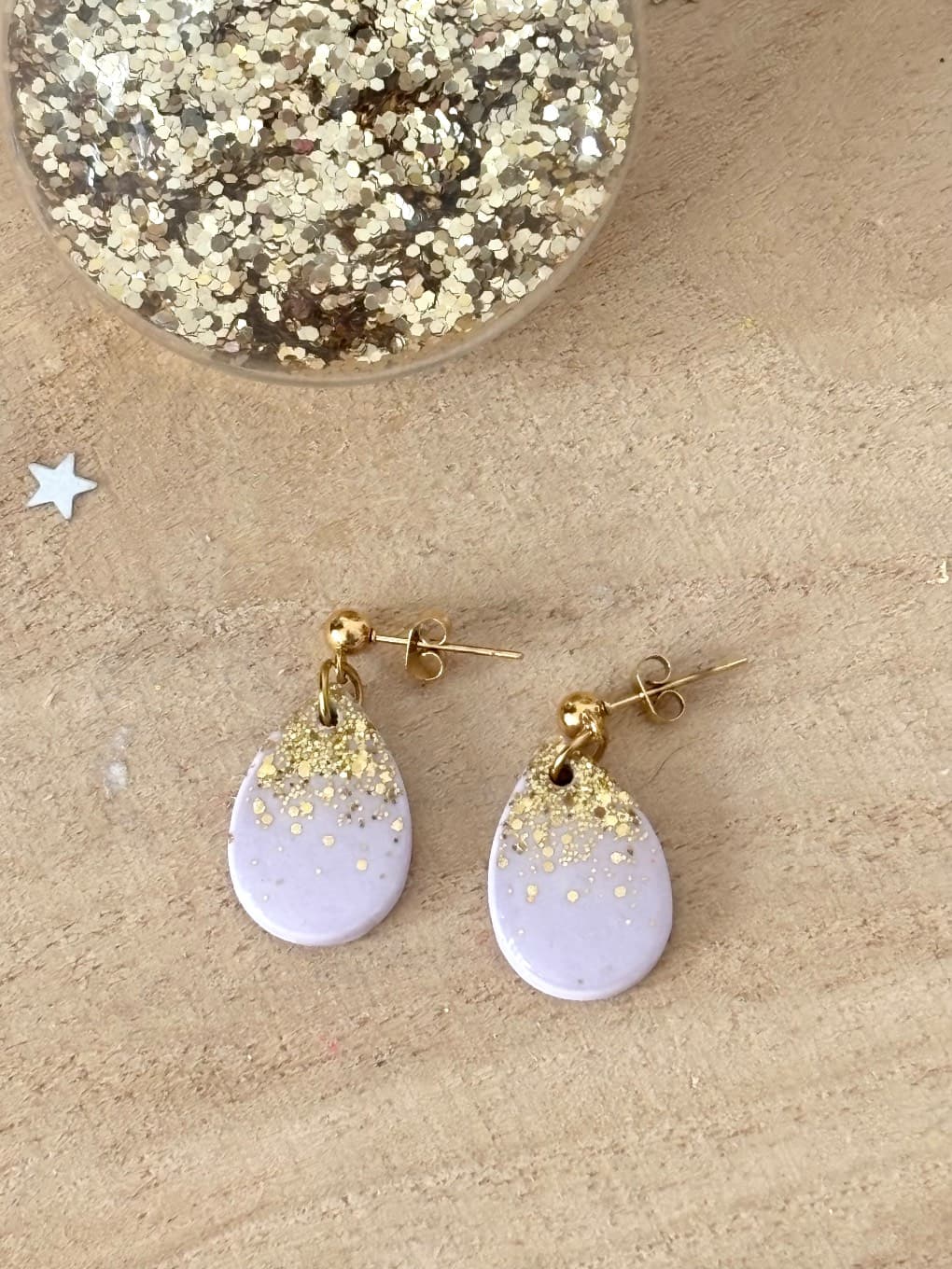 Boucles d'oreilles gouttes lilas et dégradé pailleté doré posées sur bois à côté d'un pot de paillettes