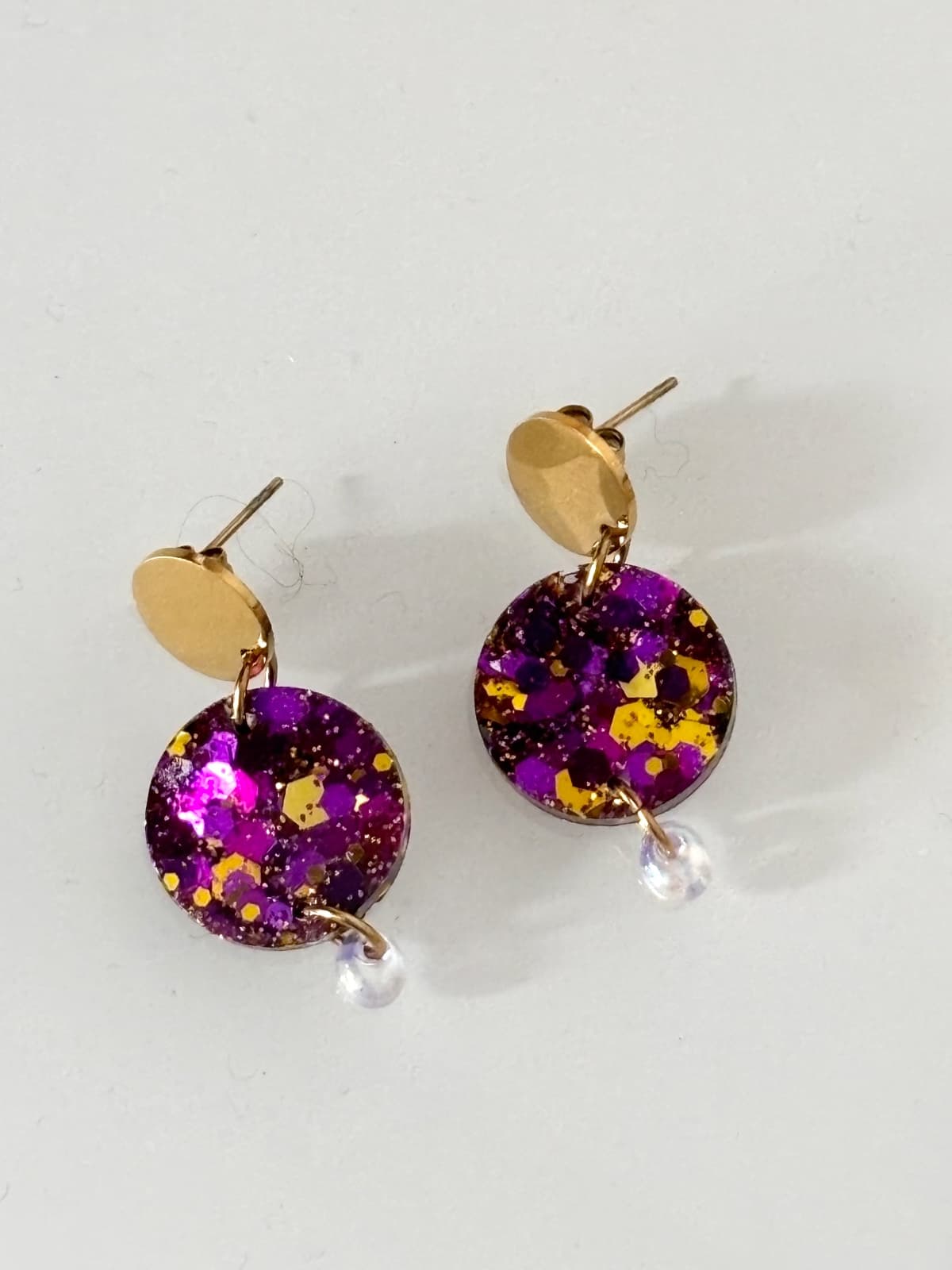 Paire de boucles d'oreilles dorées avec médaillons pailletés violet et or sur fond blanc.