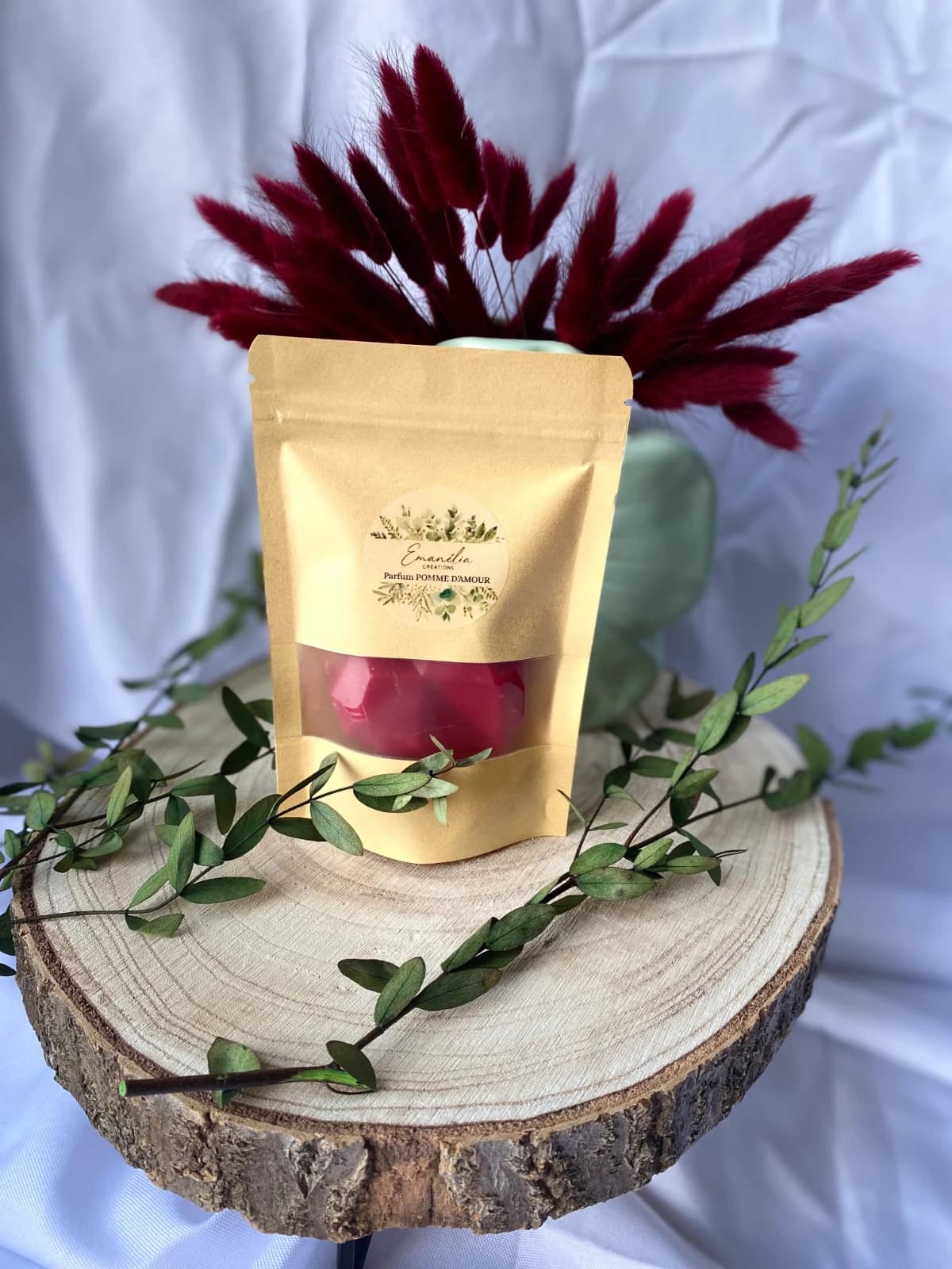 Sachet kraft avec fenêtre dévoilant un fondant rouge, posé sur un rondin de bois décoré de feuillage.