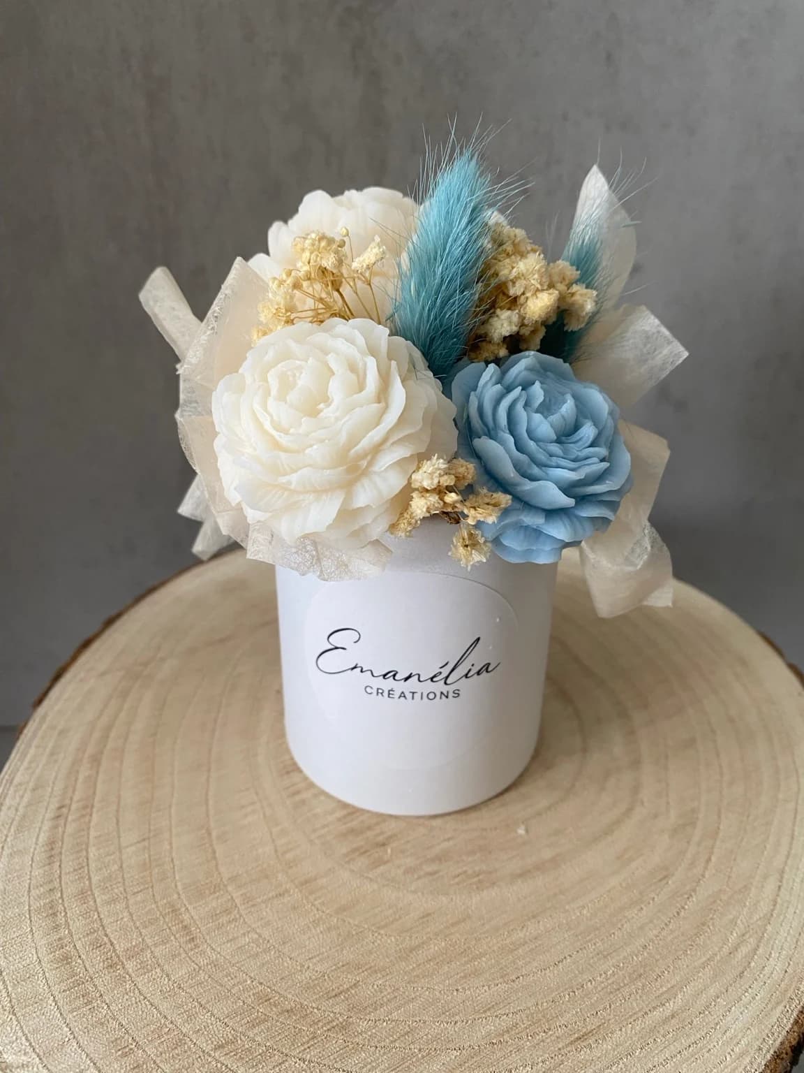 Bouquet de fleurs en cire ivoire et bleu poudré dans son pot blanc, posé sur une rondelle de bois.